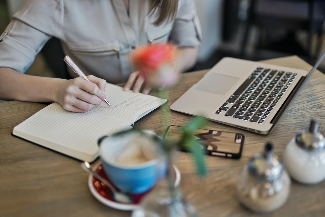 Person Writing On A Notebook Beside a laptop