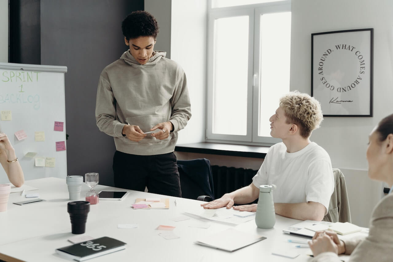 Man in Gray Sweater Standing In Front of His Colleagues Presenting